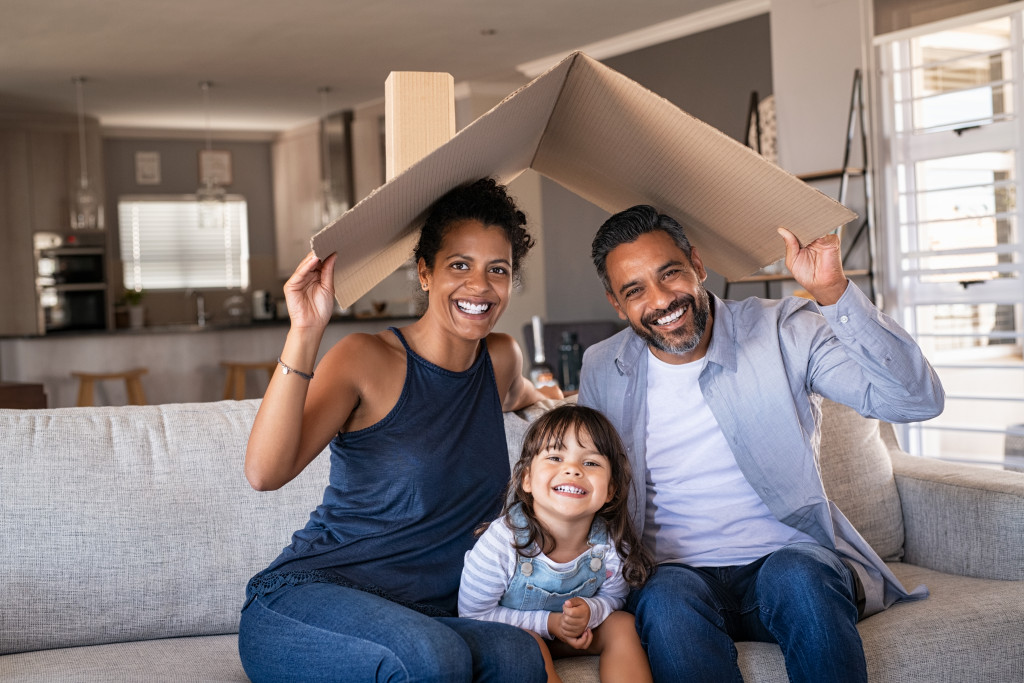 A smiling young family in the living room holding a cardboard to mimic a roof on their heads